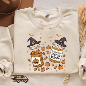 Op de afbeelding: Zandkleurige sweatshirt met een Halloween-thema. Het ontwerp toont twee medicijnflesjes met heksenhoeden, pompoenen en de tekst "Prescription: Spooky Season". Vleermuizen en sterren maken de feestelijke look compleet.