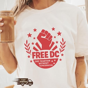 May include: Ivory-colored t-shirt with a red graphic featuring a raised fist, stars, and the text "FREE DC" and "END TAXATION WITHOUT REPRESENTATION." The shirt has short sleeves and a crew neck.
