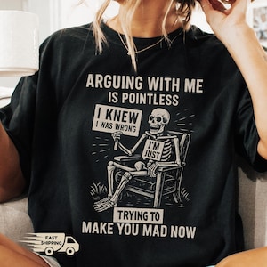 May include: Black t-shirt with a humorous skeleton graphic. The skeleton is seated in a chair and holds a sign that reads "I'M JUST". The shirt also features the text "ARGUING WITH ME IS POINTLESS" and "TRYING TO MAKE YOU MAD NOW".