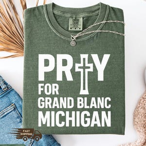 May include: A moss green t-shirt with the words "PRAY FOR GRAND BLANC MICHIGAN" in white block letters. A silver necklace with a pendant rests on the shirt. The shirt is folded and placed on a white surface.