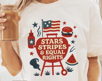 May include: Ivory-colored t-shirt featuring a red barbecue grill design with the text "STARS STRIPES & EQUAL RIGHTS." The design includes an American flag, stars, a hamburger, and watermelon slices. The shirt is a casual wear item.