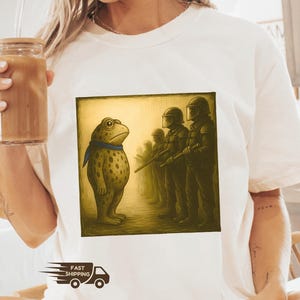 K&ouml;nnte beinhalten: Elfenbeinfarbenes T-Shirt mit einem quadratischen Aufdruck, der einen Frosch zeigt, der Soldaten gegen&uuml;bersteht. Der Frosch ist hellbraun mit dunklen Flecken und einem blauen Schal. Die Soldaten tragen dunkle Uniformen. Das T-Shirt hat einen Rundhalsausschnitt und kurze &Auml;rmel.