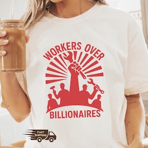 May include: Ivory t-shirt with a red graphic. The graphic shows a raised fist holding a wrench, with the words "WORKERS OVER BILLIONAIRES" in a bold font. Silhouettes of workers with tools are below the text.