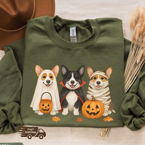 Może przedstawiać: Zielona bluza w kolorze wojskowym z trzema corgi przebranymi za Halloween. Jeden corgi jest duchem, drugi wampirem, a trzeci mumią. Bluza ma napis "Fast Shipping".