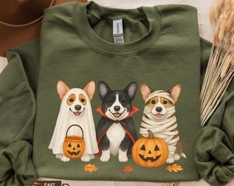 May include: Military green sweatshirt featuring three corgis dressed in Halloween costumes. One corgi is a ghost holding a jack-o'-lantern, another is a vampire, and the third is a mummy. The sweatshirt has the words "Fast Shipping" on it.