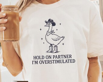 May include: Ivory-colored t-shirt featuring a cartoon goose wearing a cowboy hat, boots, and a bandana. The text "HOLD ON PARTNER I'M OVERSTIMULATED" is printed below the graphic. The shirt has a relaxed fit.