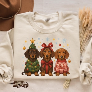 May include: A cream-colored sweatshirt featuring three dachshunds in festive holiday attire. One dog is adorned with a Christmas tree, another with a large red bow, and the third in a red sweater. The design includes snowflakes and star accents.