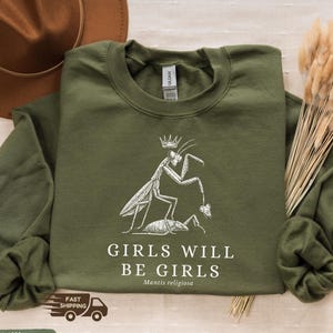 Puede incluir: Sudadera verde militar con un gráfico blanco de una mantis religiosa coronada, sosteniendo un insecto. El texto dice "GIRLS WILL BE GIRLS" y "Mantis religiosa".