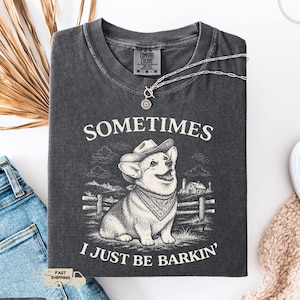 May include: Dark gray t-shirt with the text "SOMETIMES I JUST BE BARKIN'" and a graphic of a corgi wearing a cowboy hat and bandana. A silver necklace is draped across the shirt. The shirt is folded next to a pair of blue jeans.