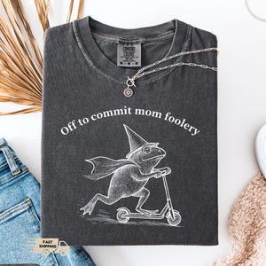May include: Dark gray t-shirt with the phrase "Off to commit mom foolery" and a graphic of a frog riding a scooter. The shirt is made of a soft material and has a crew neck. A silver necklace with a sun pendant is on the shirt.