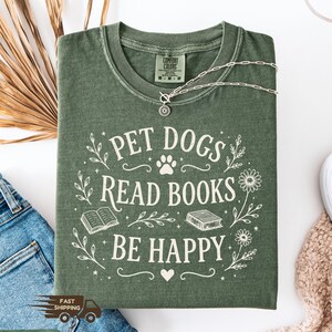 May include: A moss green t-shirt with the text "PET DOGS READ BOOKS BE HAPPY" in white script. The design includes a paw print, books, and floral accents. A silver necklace with a sun pendant is draped across the shirt. The shirt is folded on a white surface.