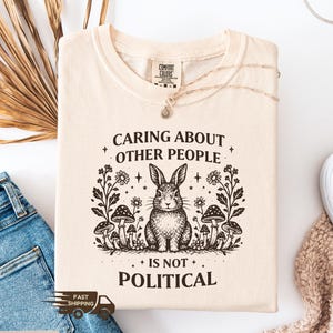 May include: Ivory-colored t-shirt with the text "Caring About Other People Is Not Political" and a graphic of a rabbit surrounded by flowers and mushrooms. The shirt is made of a soft material.