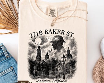 May include: Cream-coloured t-shirt with a black and white graphic of 221B Baker St, London. The design includes a silhouette of a man with a pipe, buildings, and the text "221B BAKER ST. EST. 1892".