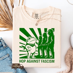 May include: Ivory-colored t-shirt with a green graphic featuring a frog, figures in riot gear, and the text "HOP AGAINST FASCISM." The design includes a sunburst pattern. The shirt is made by Comfort Colors.