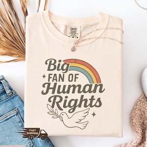以下が含まれることがあります： 「Big Fan of Human Rights」の文字がレトロなフォントでプリントされたアイボリー色のTシャツ。虹とオリーブの枝を持った鳩が描かれています。Tシャツは折りたたまれており、ネックレスとジーンズが見えます。