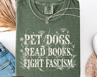 May include: A moss green t-shirt with the text "PET DOGS READ BOOKS FIGHT FASCISM" in white, surrounded by floral illustrations. A silver necklace with a sunburst pendant rests on the shirt. The shirt is folded on a white surface with denim jeans and a beige sweater.