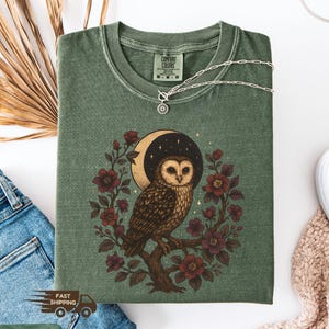 May include: A moss green t-shirt with an owl design. The illustration shows a brown owl on a branch, encircled by burgundy flowers and a crescent moon. A silver necklace is placed on the shirt. The shirt is folded.