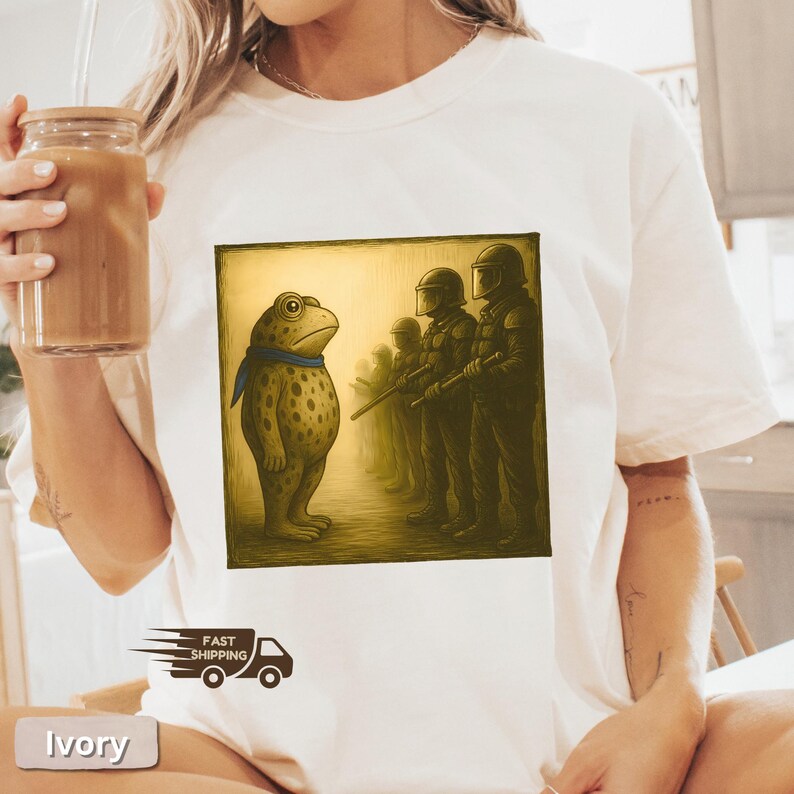 K&ouml;nnte beinhalten: Elfenbeinfarbenes T-Shirt mit einem quadratischen Aufdruck, der einen Frosch zeigt, der Soldaten gegen&uuml;bersteht. Der Frosch ist hellbraun mit dunklen Flecken und einem blauen Schal. Die Soldaten tragen dunkle Uniformen. Das T-Shirt hat einen Rundhalsausschnitt und kurze &Auml;rmel.