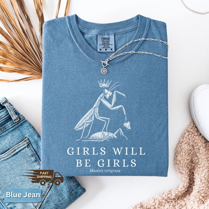 May include: Blue jean-colored t-shirt with a crew neck and short sleeves. The shirt features a white graphic of a praying mantis wearing a crown, with the text "GIRLS WILL BE GIRLS" and "Mantis religiosa" below. A silver necklace rests on the shirt.