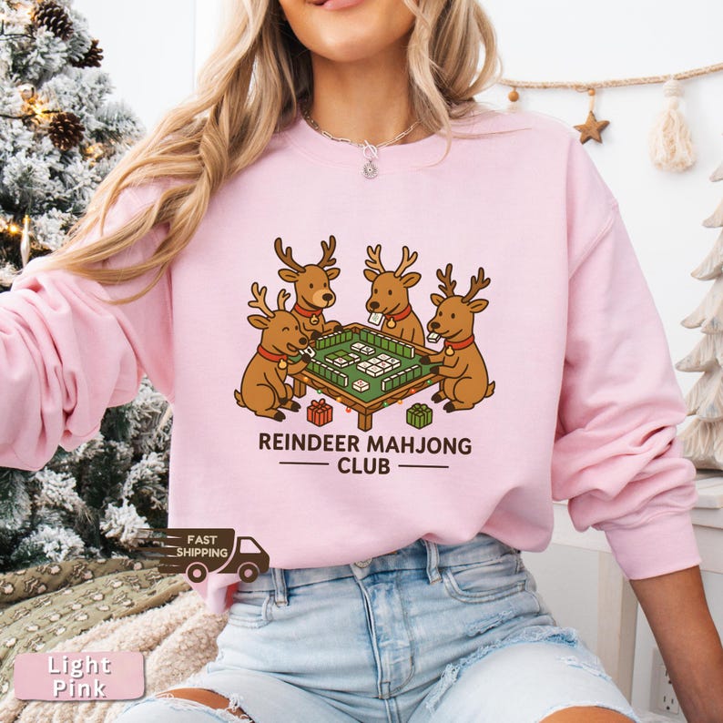 Puede incluir: Sudadera rosa claro con una ilustraci&oacute;n de dibujos animados de cuatro renos jugando al mahjong. Los renos marrones con cuernos est&aacute;n reunidos alrededor de una mesa de mahjong verde. El texto "REINDEER MAHJONG CLUB" est&aacute; impreso debajo.
