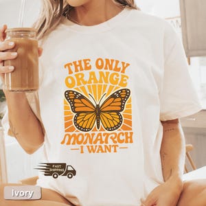 Op de afbeelding: Ivoor kleurig T-shirt met de tekst "THE ONLY ORANGE MONARCH I WANT" in oranje. Het ontwerp toont een oranje en zwarte monarchvlinder met een zonnestralen achtergrond. Het shirt heeft een casual stijl.