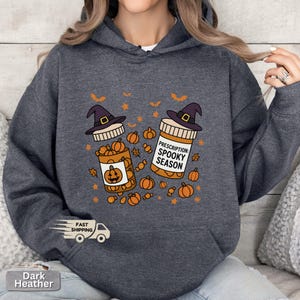 Op de afbeelding: Donkergrijze hoodie met een Halloween-ontwerp. De afbeelding toont twee pillenflesjes met heksenhoeden, pompoenen en de tekst "Prescription: Spooky Season". De hoodie heeft een voorzak en een capuchon met trekkoord.