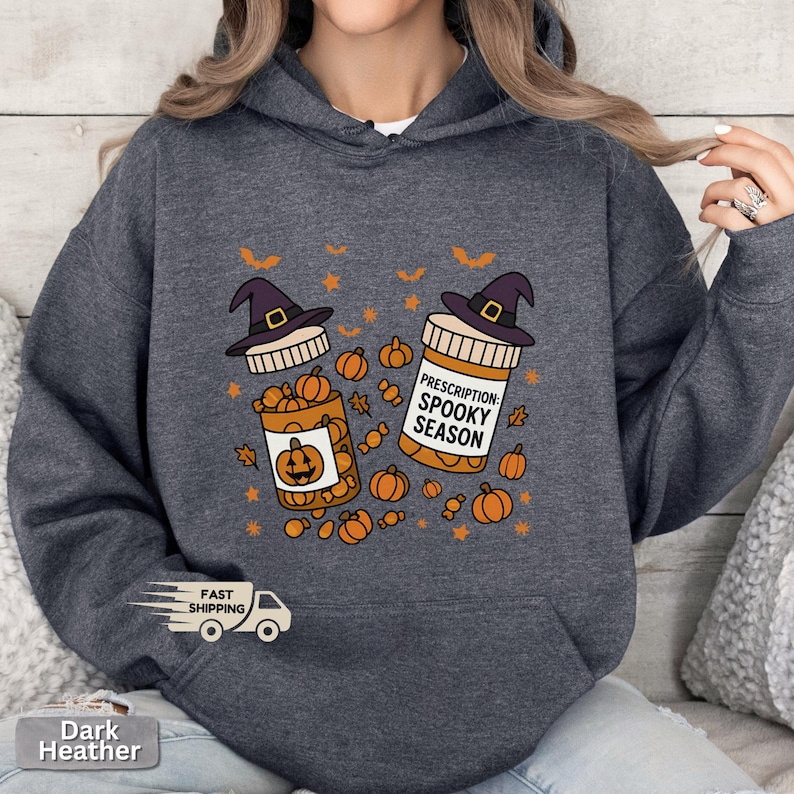 Op de afbeelding: Donkergrijze hoodie met een Halloween-ontwerp. De afbeelding toont twee pillenflesjes met heksenhoeden, pompoenen en de tekst "Prescription: Spooky Season". De hoodie heeft een voorzak en een capuchon met trekkoord.