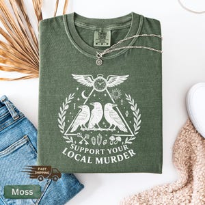 May include: A moss green t-shirt with a white graphic featuring three crows, a clock with wings, and the text "SUPPORT YOUR LOCAL MURDER." A silver necklace with a pendant rests on the shirt. The shirt is folded on a white surface.