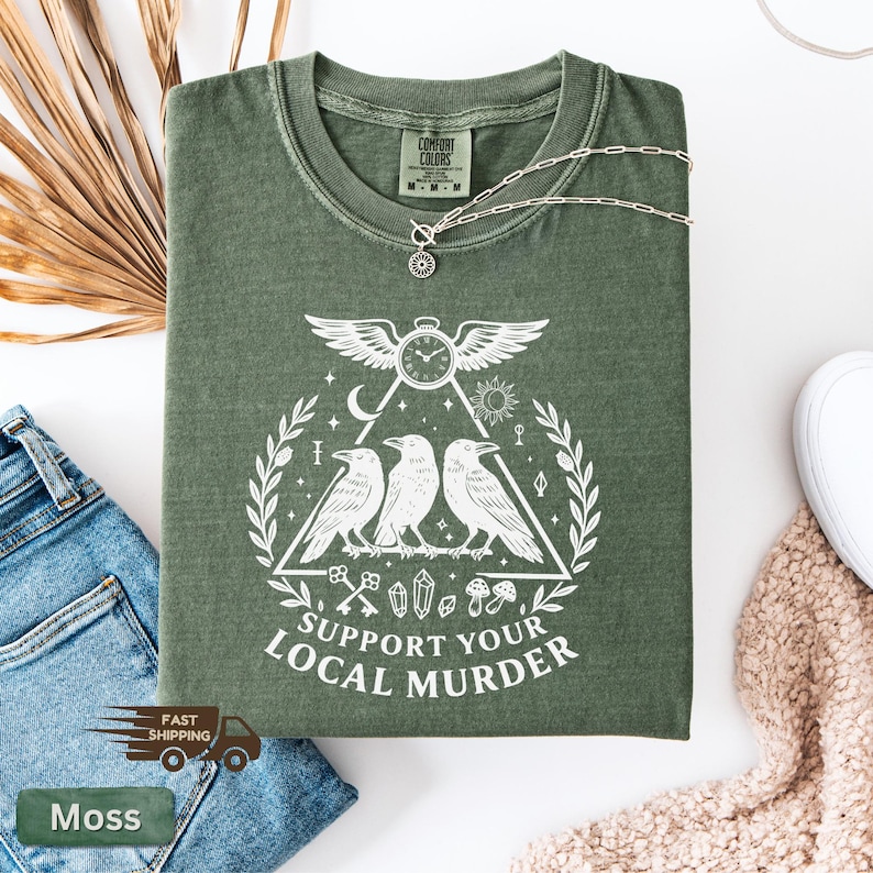 May include: A moss green t-shirt with a white graphic featuring three crows, a clock with wings, and the text "SUPPORT YOUR LOCAL MURDER." A silver necklace with a pendant rests on the shirt. The shirt is folded on a white surface.