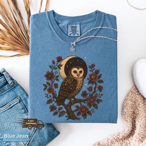 May include: Blue jean coloured t-shirt with a crew neck. The shirt features a detailed illustration of an owl perched on a branch, surrounded by flowers and a crescent moon. A silver necklace with a sun pendant rests on the shirt. The words "Comfort Colors" are printed on the shirt.