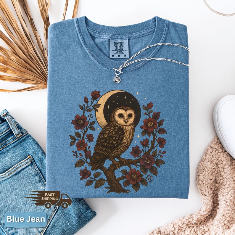 May include: Blue jean coloured t-shirt with a crew neck. The shirt features a detailed illustration of an owl perched on a branch, surrounded by flowers and a crescent moon. A silver necklace with a sun pendant rests on the shirt. The words "Comfort Colors" are printed on the shirt.