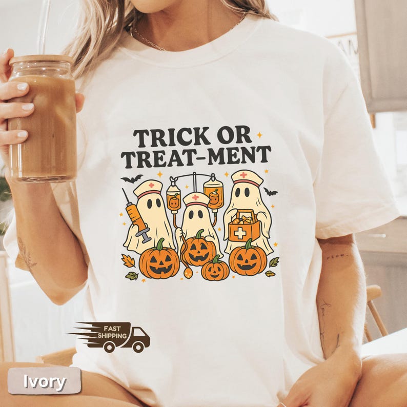 May include: Ivory-colored t-shirt with a Halloween-themed graphic. The design features cartoon ghosts dressed as nurses, pumpkins, and the text "TRICK OR TREAT-MENT". The shirt has a crew neck and short sleeves.