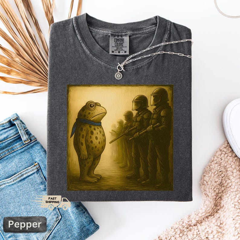 K&ouml;nnte beinhalten: Dunkelgraues Comfort Colors T-Shirt mit einer humorvollen Illustration einer Froschfigur, die sich Soldaten gegen&uuml;bersieht. Der Frosch tr&auml;gt einen blauen Schal. Eine silberne Halskette mit Anh&auml;nger ist ebenfalls sichtbar. Das Shirt ist mit "Pepper" beschriftet.