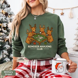 Puede incluir: Sudadera verde militar con un dise&ntilde;o de cuatro renos jugando al mahjong. El texto "REINDEER MAHJONG CLUB" est&aacute; debajo del dise&ntilde;o. La persona lleva un pantal&oacute;n de pijama a cuadros rojos y blancos y sostiene una taza blanca con un dise&ntilde;o de Pap&aacute; Noel.