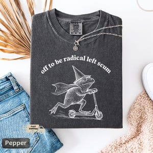 Pode incluir: T-shirt cinza escuro com o texto "off to be radical left scum" e um gr&aacute;fico de um sapo andando de scooter, usando um chap&eacute;u de mago e capa. A camisa est&aacute; dobrada em uma superf&iacute;cie branca.