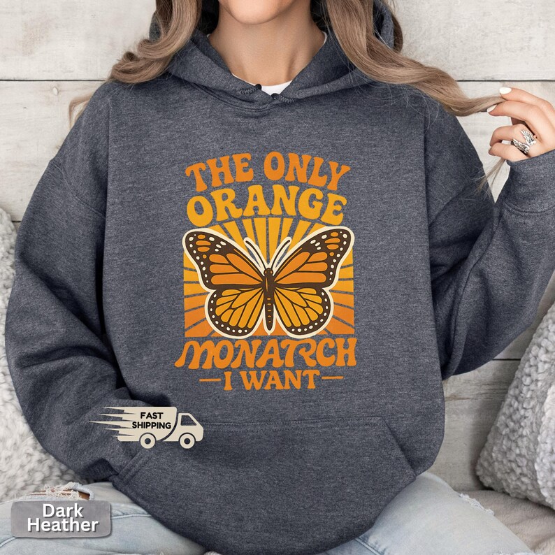 Op de afbeelding: Donkergrijze hoodie met de tekst "THE ONLY ORANGE MONARCH I WANT" in oranje. Een monarchvlinder-afbeelding staat in het midden, met oranje en gele stralen erachter. Een casual kledingstuk.