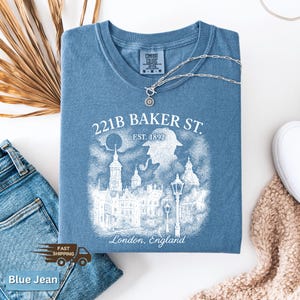 May include: A blue t-shirt with a graphic that reads "221B BAKER ST. EST. 1892 London, England". The graphic depicts a silhouette of a person with a pipe, buildings, and street lamps. A silver necklace is draped across the shirt.