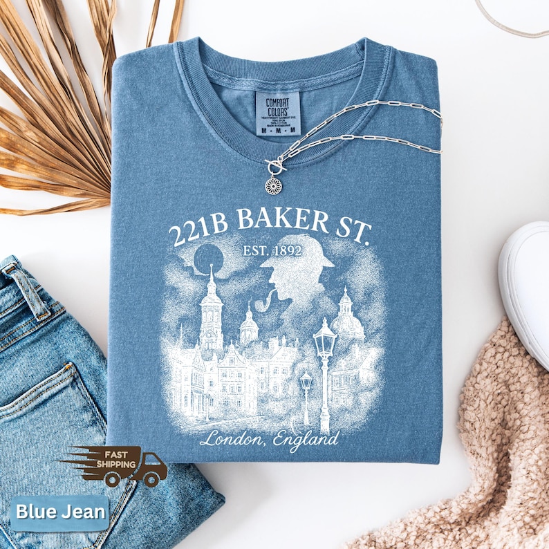 May include: A blue t-shirt with a graphic that reads "221B BAKER ST. EST. 1892 London, England". The graphic depicts a silhouette of a person with a pipe, buildings, and street lamps. A silver necklace is draped across the shirt.