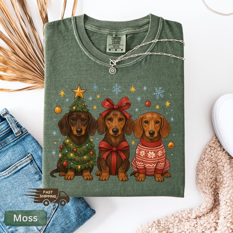 May include: Moss green Comfort Colors t-shirt featuring three dachshunds. One is decorated as a Christmas tree, another with a red bow, and the third in a red and white sweater. The shirt has a silver necklace and the text "Moss".
