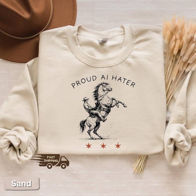 May include: Sand-colored sweatshirt with the text "PROUD AI HATER" above a graphic of a cowboy riding a rearing horse. Below the horse are three red stars. A brown hat and dried plants are in the background.