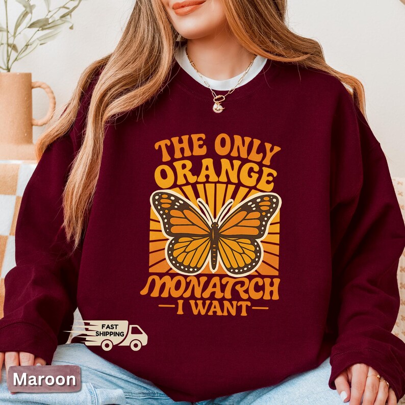 Op de afbeelding: Bordeaux sweatshirt met een afbeelding van een monarchvlinder en de tekst "THE ONLY ORANGE MONARCH I WANT". De vlinder is oranje en zwart, tegen een gele en oranje achtergrond.