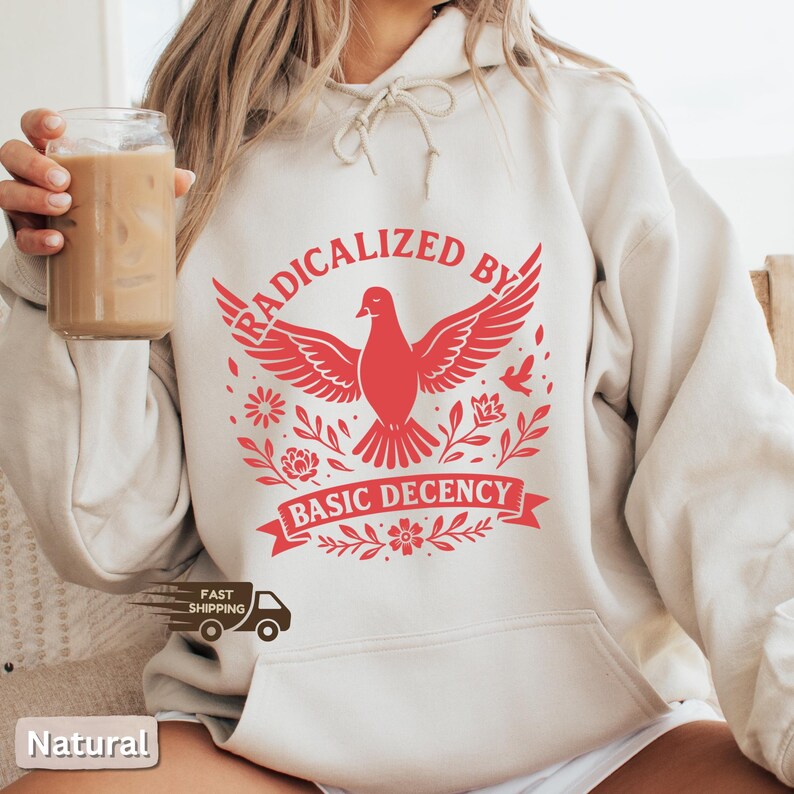 Op de afbeelding: Een cr&egrave;mekleurige hoodie met een rode afbeelding van een duif met uitgespreide vleugels, omringd door bloemmotieven. De tekst "RADICALIZED BY" staat boven de duif en "BASIC DECENCY" eronder. De hoodie heeft een capuchon met trekkoord.