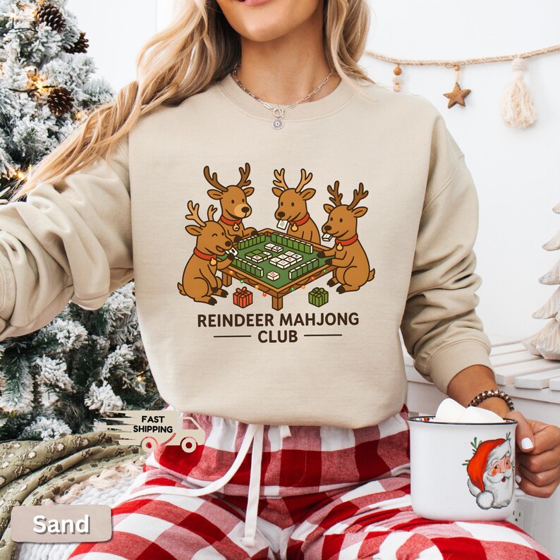 Puede incluir: Sudadera color arena con un gr&aacute;fico de cuatro renos jugando al mahjong. El texto "REINDEER MAHJONG CLUB" est&aacute; debajo de la ilustraci&oacute;n. Parte de una colecci&oacute;n tem&aacute;tica navide&ntilde;a.