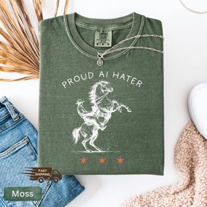 May include: Moss green t-shirt with the text "PROUD AI HATER" and a white graphic of a cowboy riding a rearing horse. The shirt has a crew neck and short sleeves. A silver necklace with a sun pendant is on top of the shirt.
