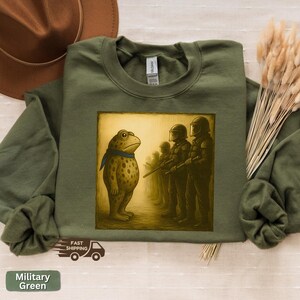 K&ouml;nnte beinhalten: Milit&auml;rgr&uuml;nes Sweatshirt mit einem Frosch-Cartoon, der einer Reihe von Soldaten gegen&uuml;bersteht. Der Frosch tr&auml;gt einen blauen Schal. Die Soldaten tragen Helme und Gewehre. Auf dem Sweatshirt steht "Military Green".