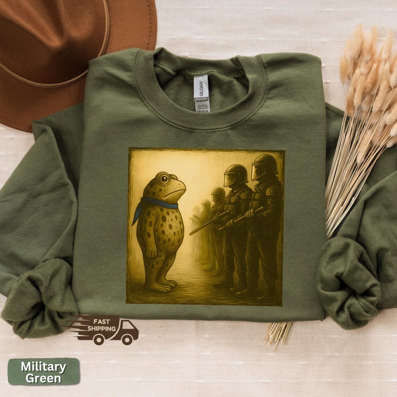 K&ouml;nnte beinhalten: Milit&auml;rgr&uuml;nes Sweatshirt mit einem Frosch-Cartoon, der einer Reihe von Soldaten gegen&uuml;bersteht. Der Frosch tr&auml;gt einen blauen Schal. Die Soldaten tragen Helme und Gewehre. Auf dem Sweatshirt steht "Military Green".