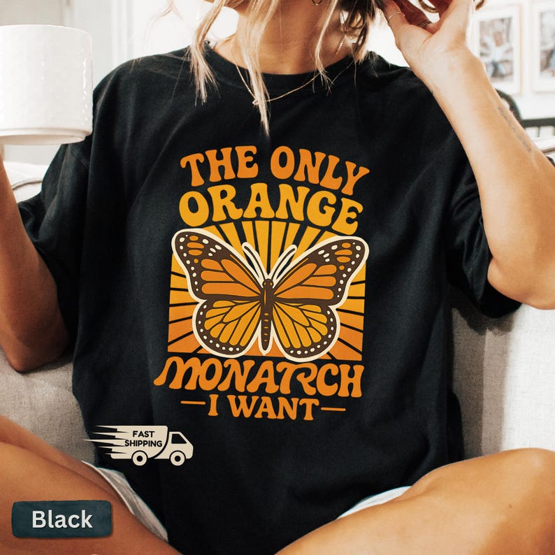 Op de afbeelding: Zwart T-shirt met de tekst "THE ONLY ORANGE MONARCH - I WANT -" in oranje en wit. Het ontwerp toont een monarchvlinder in oranje en bruin. Het shirt heeft ook een "FAST SHIPPING" afbeelding.