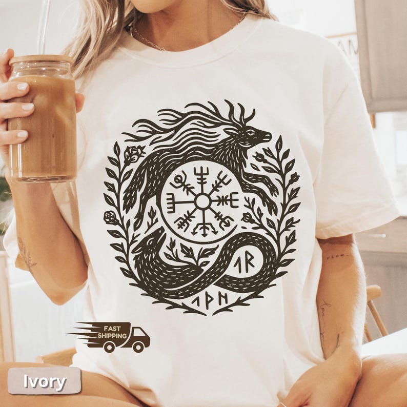 以下が含まれることがあります： アイボリー色のTシャツ。黒いグラフィックで、鹿、円形のシンボル、蛇が葉で囲まれています。Tシャツは半袖でゆったりとしたフィット感です。「Fast Shipping」の文字がトラックのアイコンとともに表示されています。