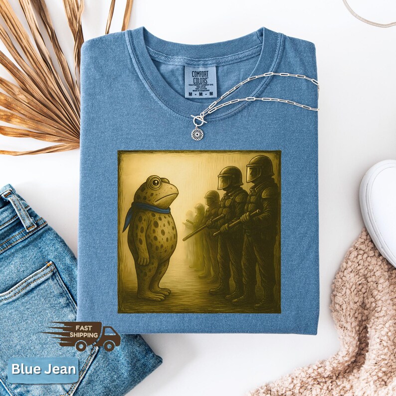 K&ouml;nnte beinhalten: Ein blaues Comfort Colors T-Shirt mit einer Grafik einer Froschfigur, die Soldaten gegen&uuml;bersteht. Der Frosch tr&auml;gt einen blauen Schal. Eine silberne Halskette mit Sonnenanh&auml;nger befindet sich auf dem Shirt. Der Text "Blue Jean" steht unten.