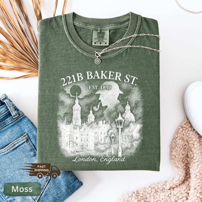 May include: A moss green t-shirt with a graphic featuring the text "221B BAKER ST. EST. 1892 London, England." The design includes an illustration of a silhouette of a person with a pipe, buildings, and street lamps. A silver necklace is draped across the shirt.